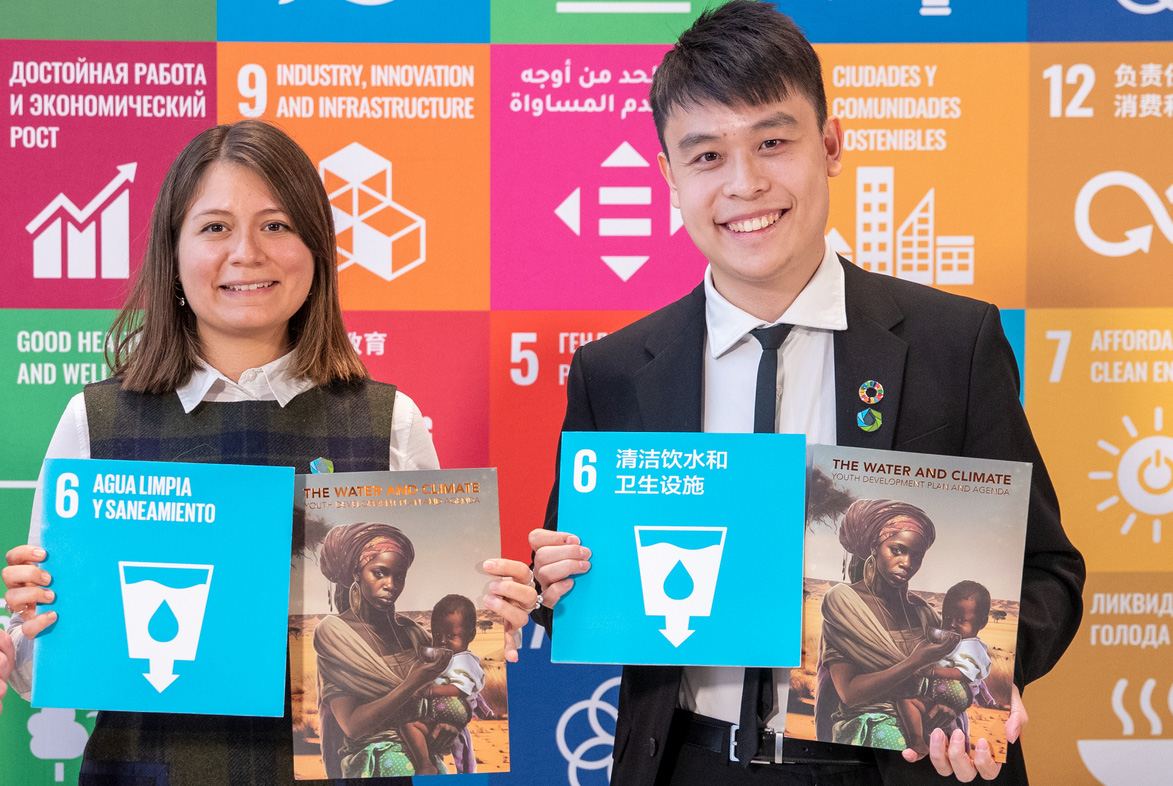 Une jeune femme et un jeune homme tenant un livre sur l'eau devant un mur montrant les cartes des 17 objectifs de développement durable dans plusieurs langues